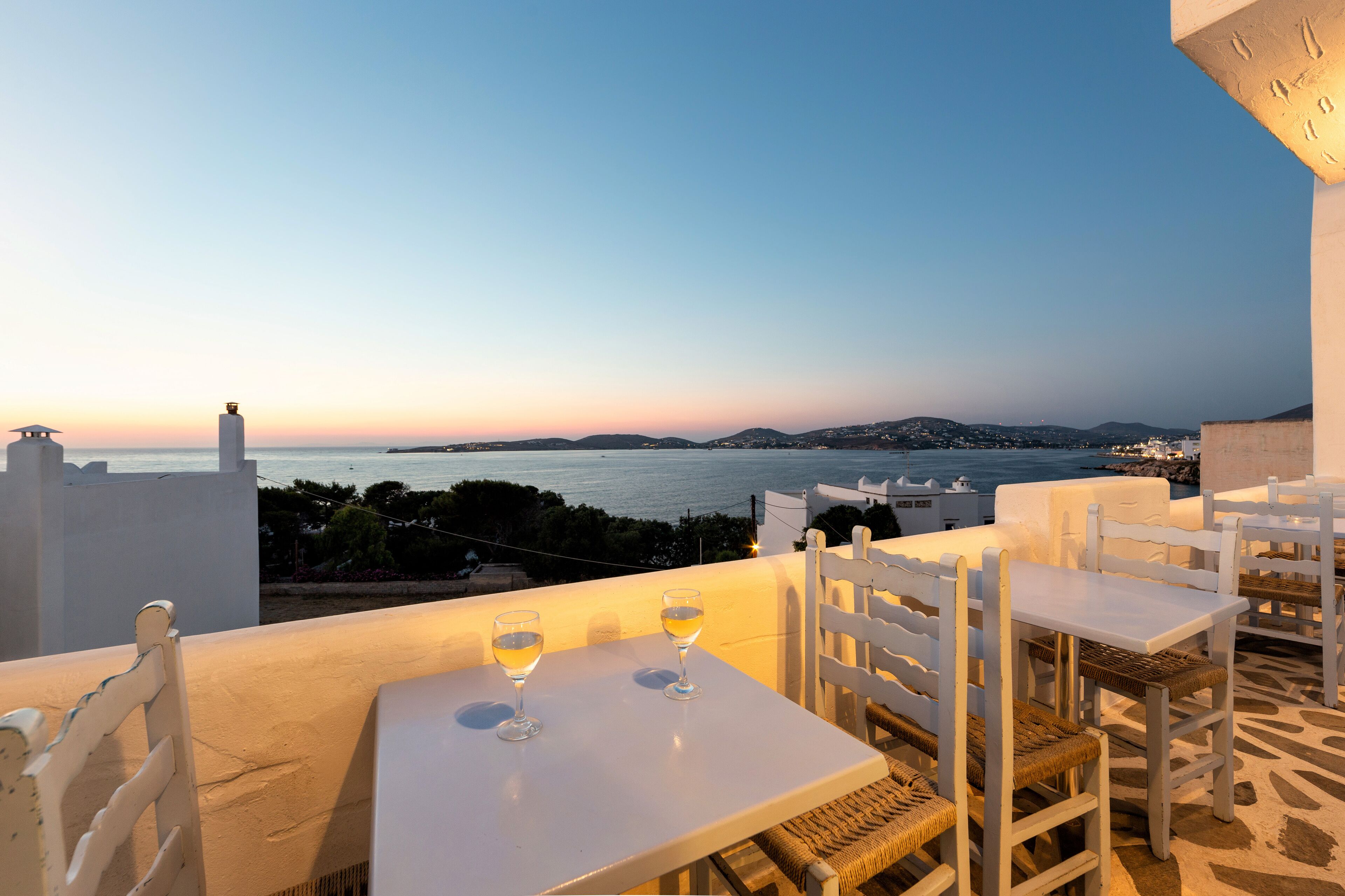 A terrace with white furniture and two glasses of wine, overlooking a coastal view with buildings and the sea.