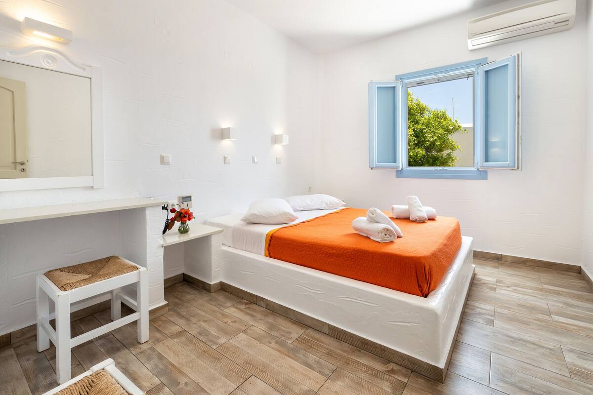 Un dormitorio moderno con una cama blanca, una colcha naranja, un banco de madera, una mesita de noche blanca, un espejo y una ventana con marcos azules.
