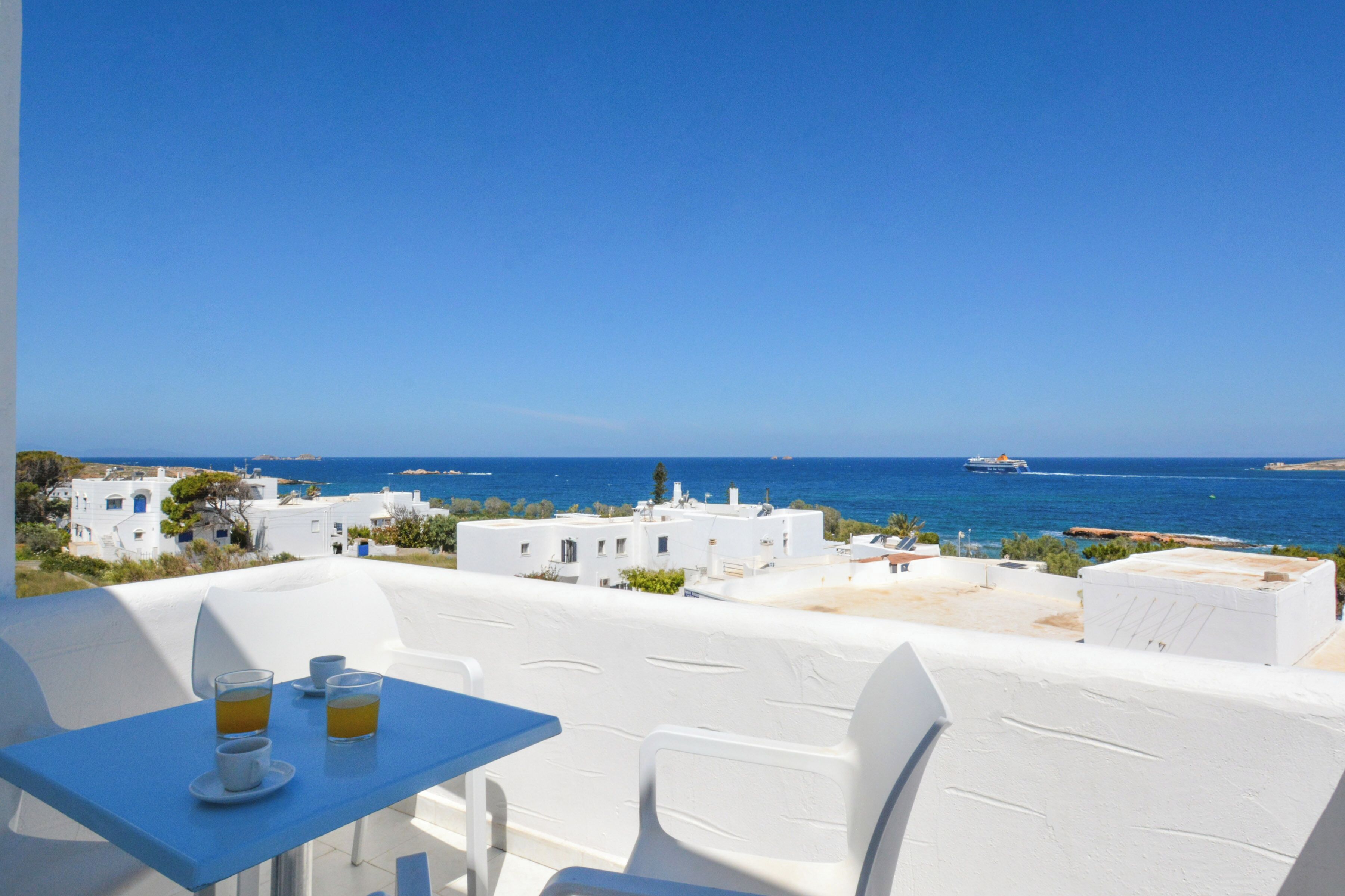 Un balcon avec des chaises blanches et une table, offrant une vue sur la côte, avec des bâtiments et la mer.
