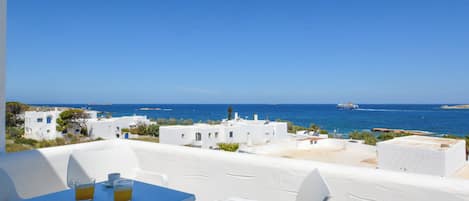 Un balcon avec des chaises blanches et une table, offrant une vue sur la côte, avec des bâtiments et la mer.