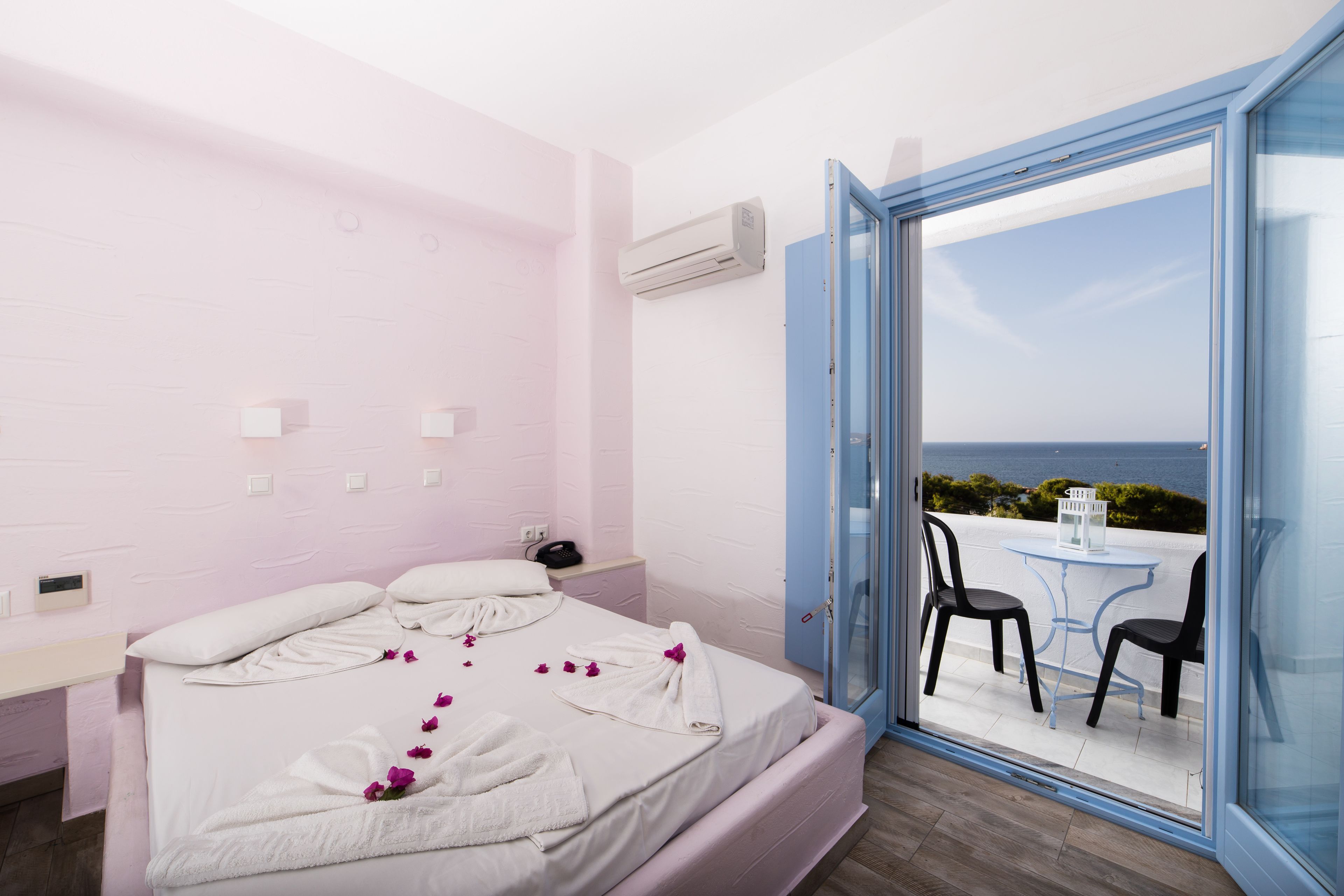 A hotel room with a bed, a nightstand, an air conditioning unit, and a balcony with a table and chairs.