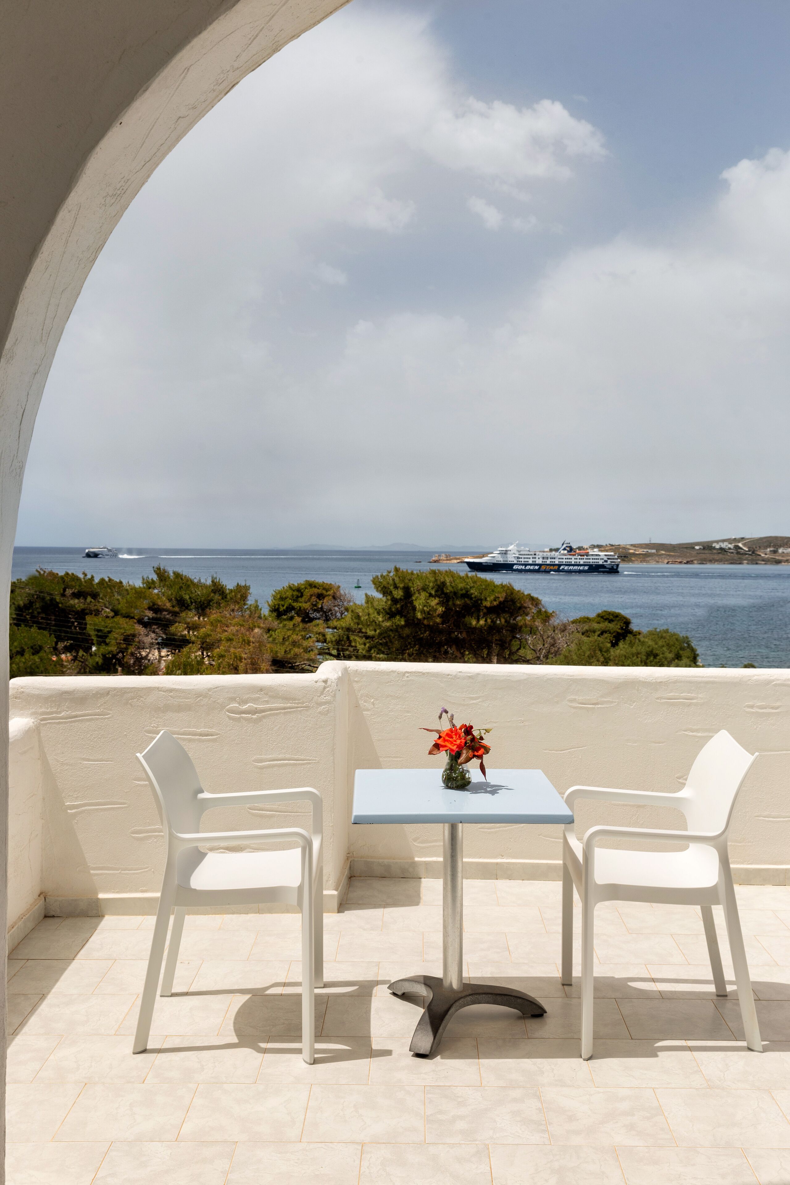 Un balcon avec deux chaises blanches et une table, donnant sur une étendue d’eau où l’on aperçoit, au loin, un navire de croisière.
