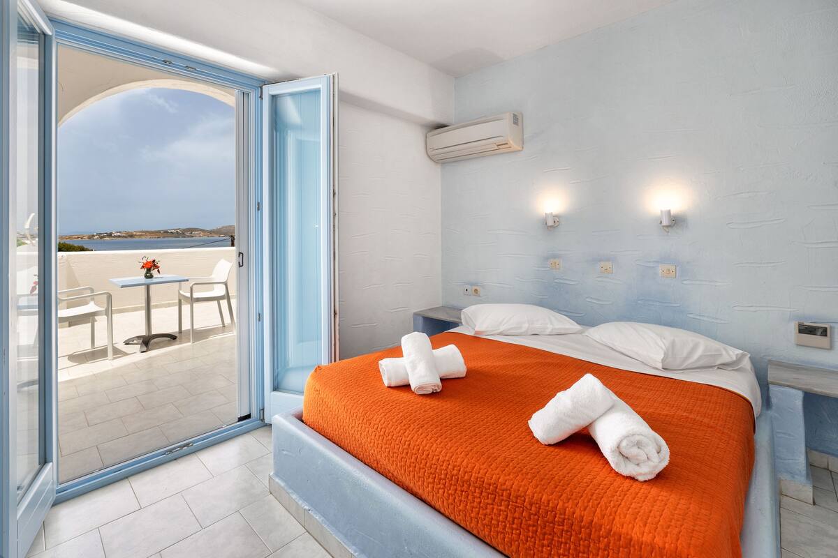 Habitación de hotel con cama, mesita de noche, ventana con vistas al mar y una mesa con sillas afuera.