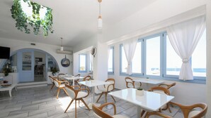 Una habitación luminosa con paredes blancas, ventanas amplias y vistas al mar. Hay mesas y sillas dispuestas para comer.