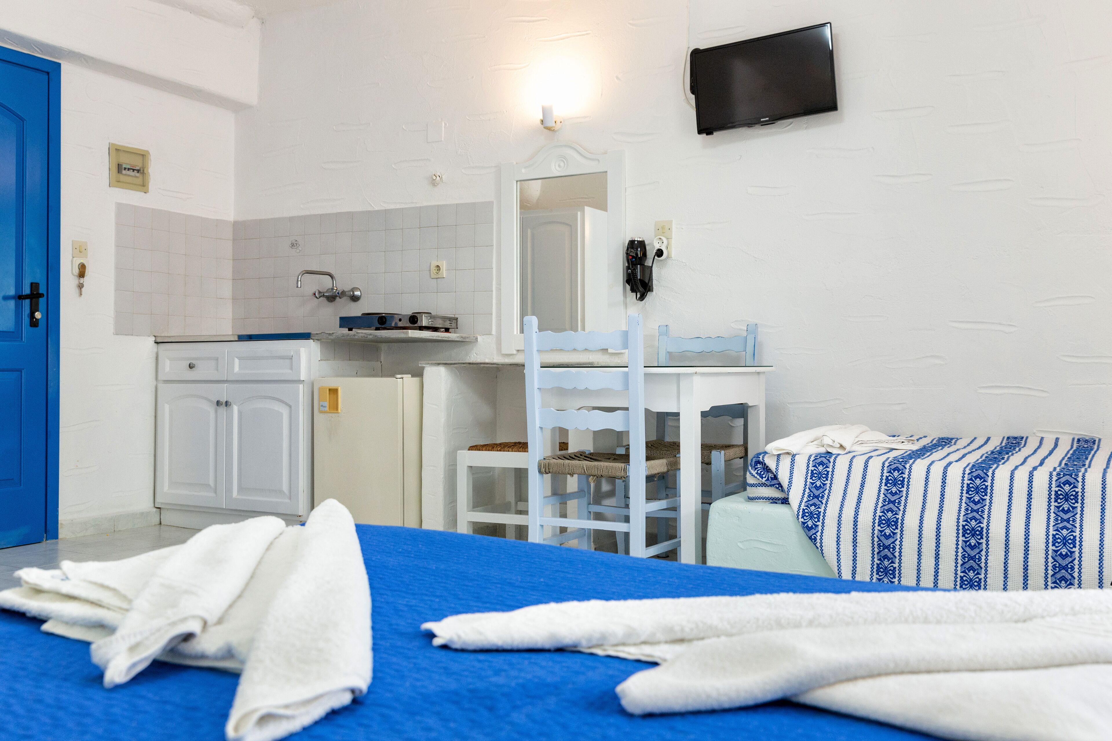 Habitación de hotel con cama, escritorio, silla, kitchenette pequeña y una puerta azul.