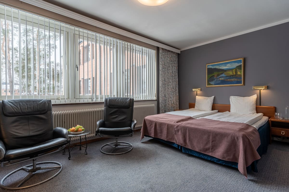 A hotel room with a bed, two chairs, a small table with fruit, a radiator, and a painting on the wall.