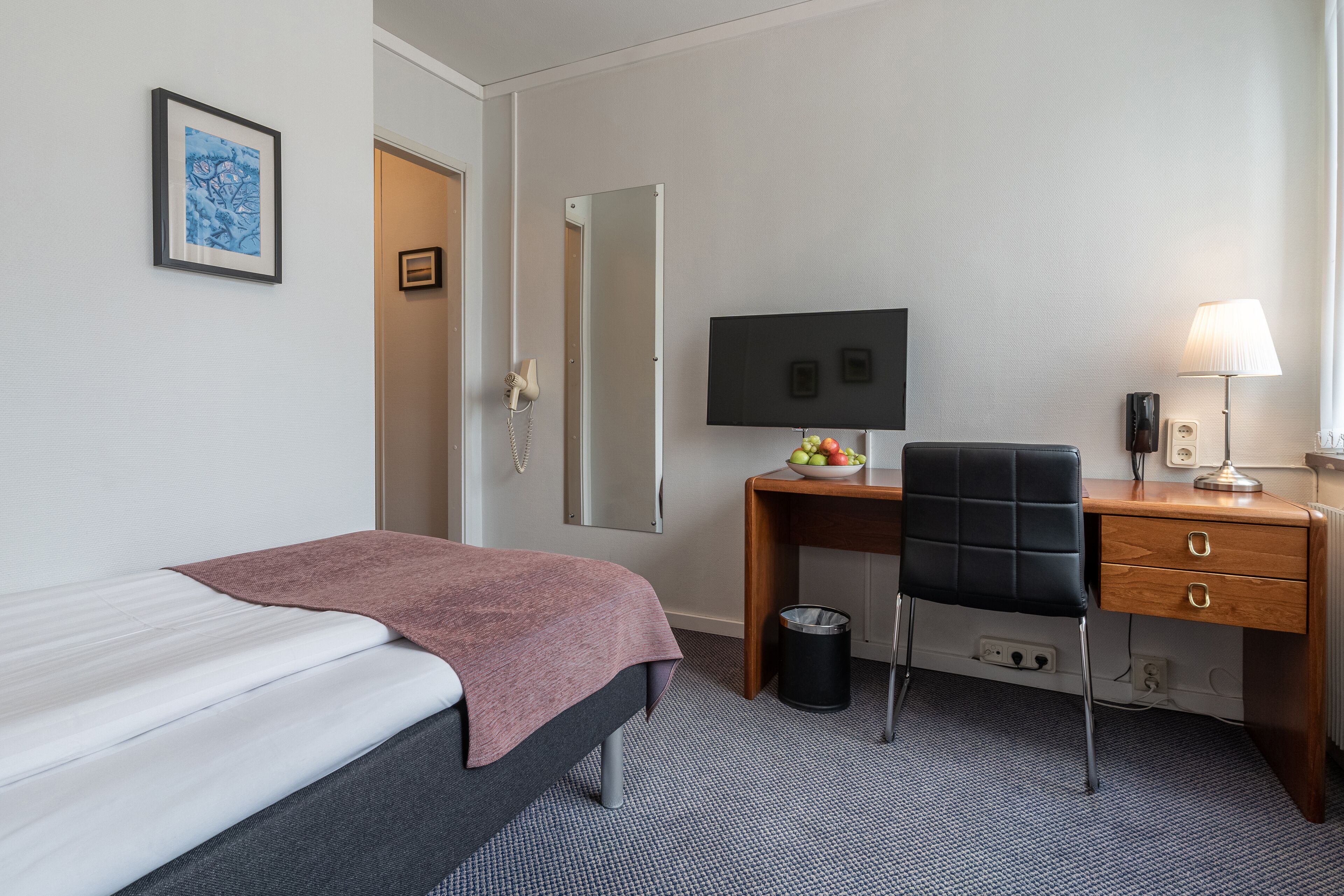 A hotel room with a bed, a desk with a computer, a chair, a trash bin, and a lamp.