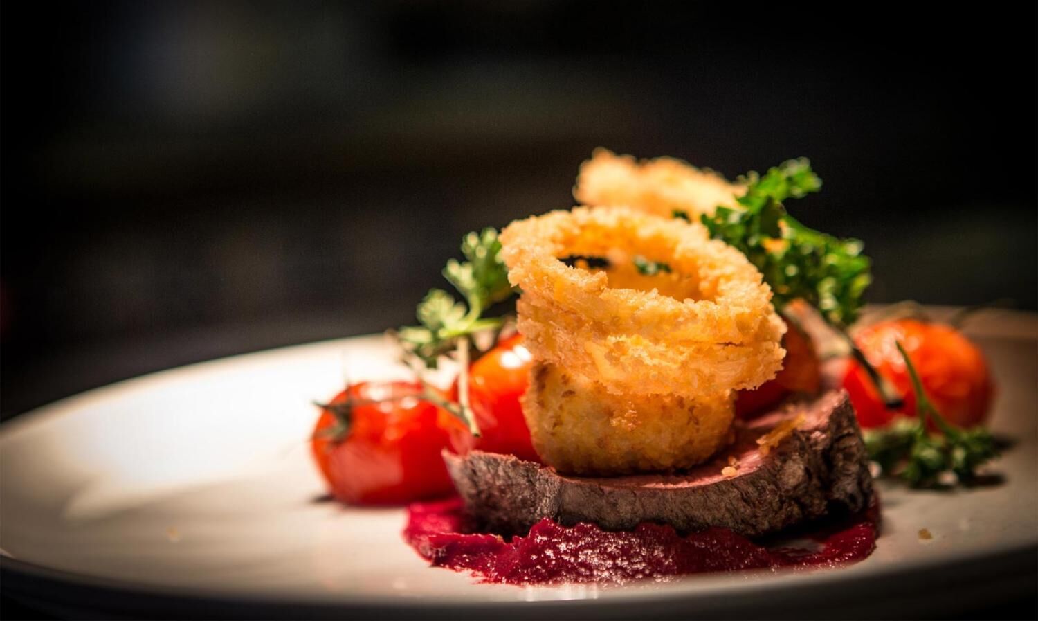 Een bord met vlees, kersen-tomaten en een gefrituurd gerecht, mogelijk een kroket, op een wit bord tegen een donkere achtergrond.