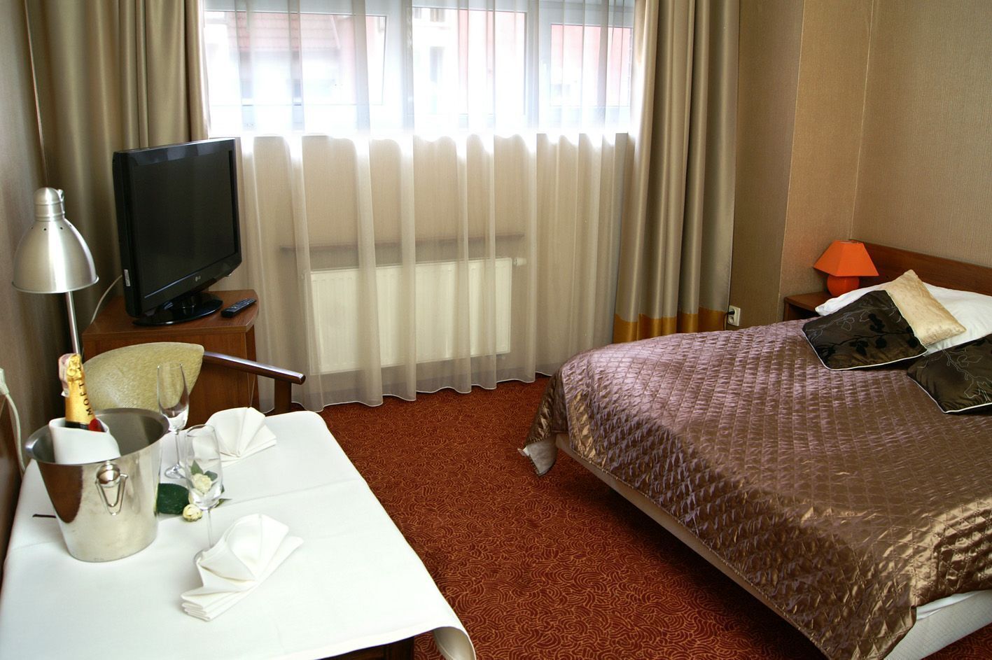 Une chambre d’hôtel avec un lit, un bureau équipé d’un ordinateur, une télévision, une lampe et une bouteille de champagne.