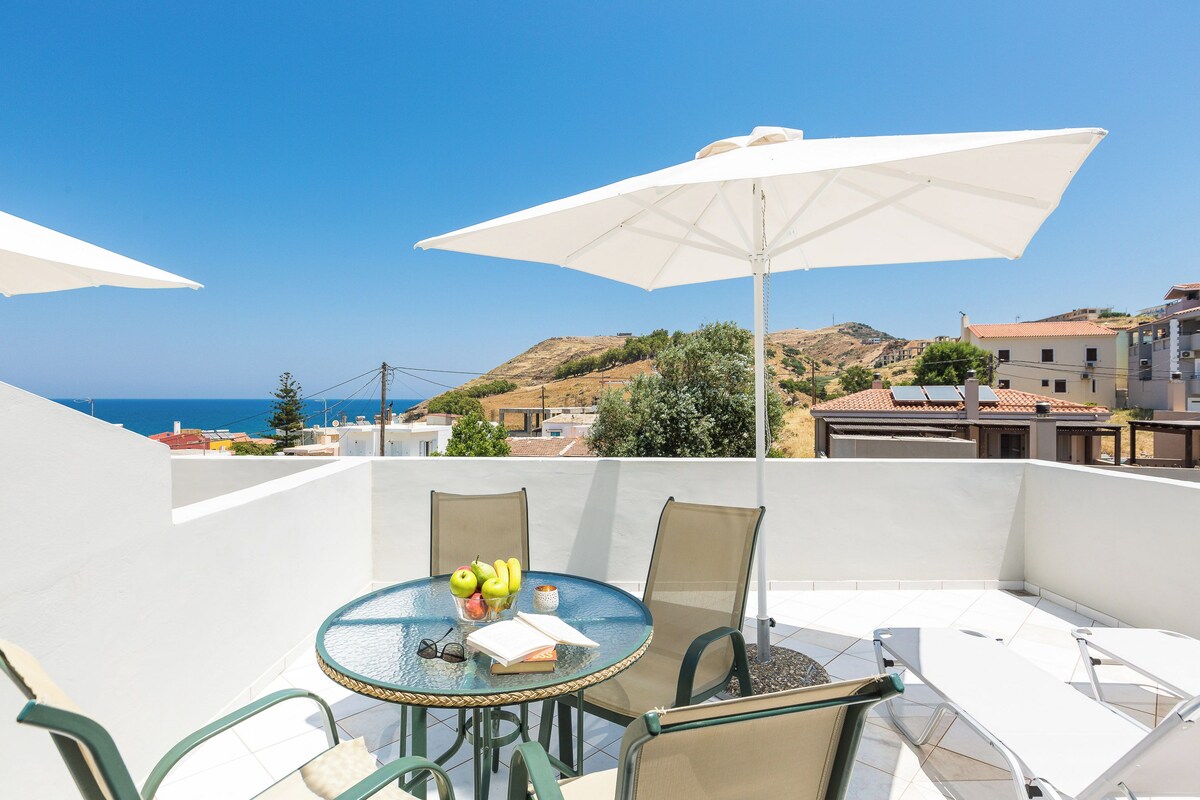 A round table with chairs and a white umbrella, overlooking a coastal view with buildings and hills.