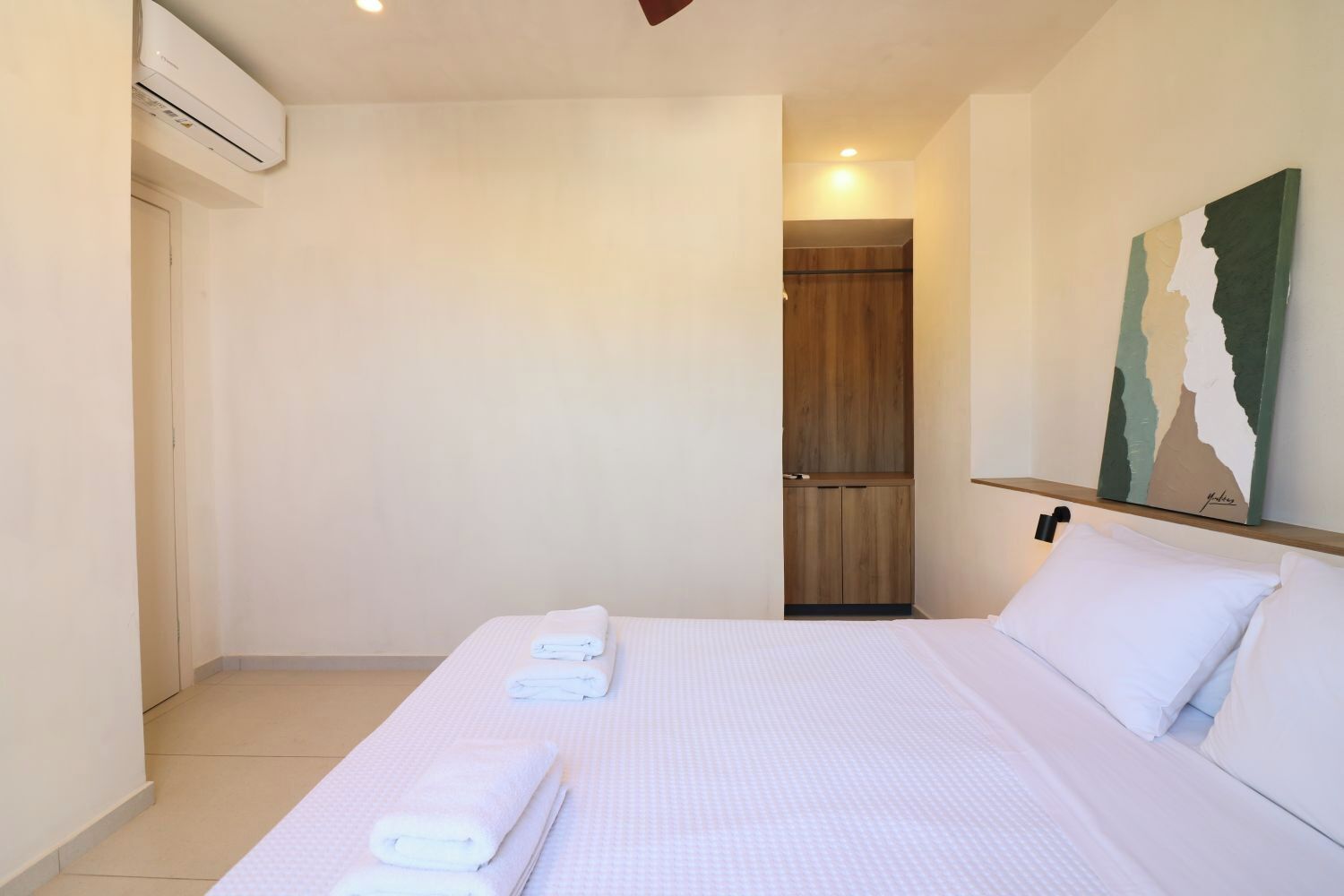 Una habitación de hotel con una cama, un armario de madera, un estante en la pared con un cuadro y un equipo de aire acondicionado.