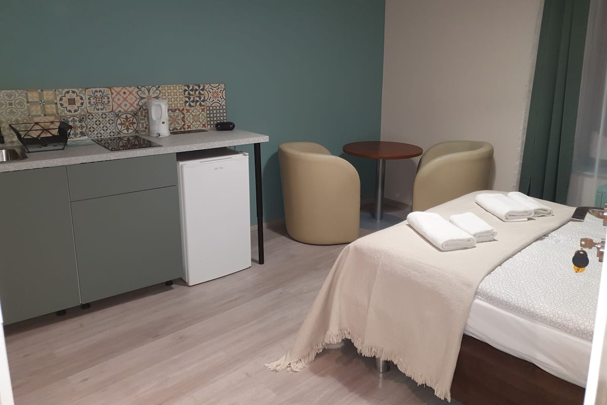 A compact hotel room with a kitchenette, a bed with a white duvet, two chairs, and a small round table.