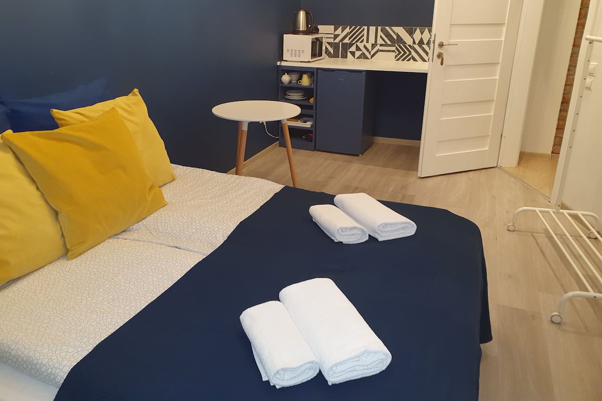 A hotel room with a bed, two pillows, a small table, a desk with a kettle, and a door.