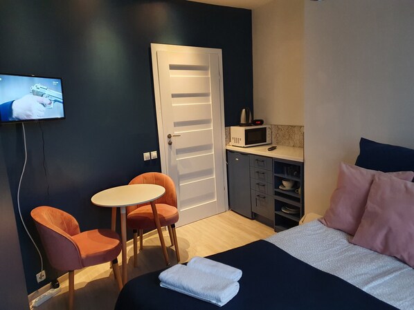 Una habitación pequeña y moderna con una cama, un escritorio con microondas, dos sillas y un televisor en la pared.