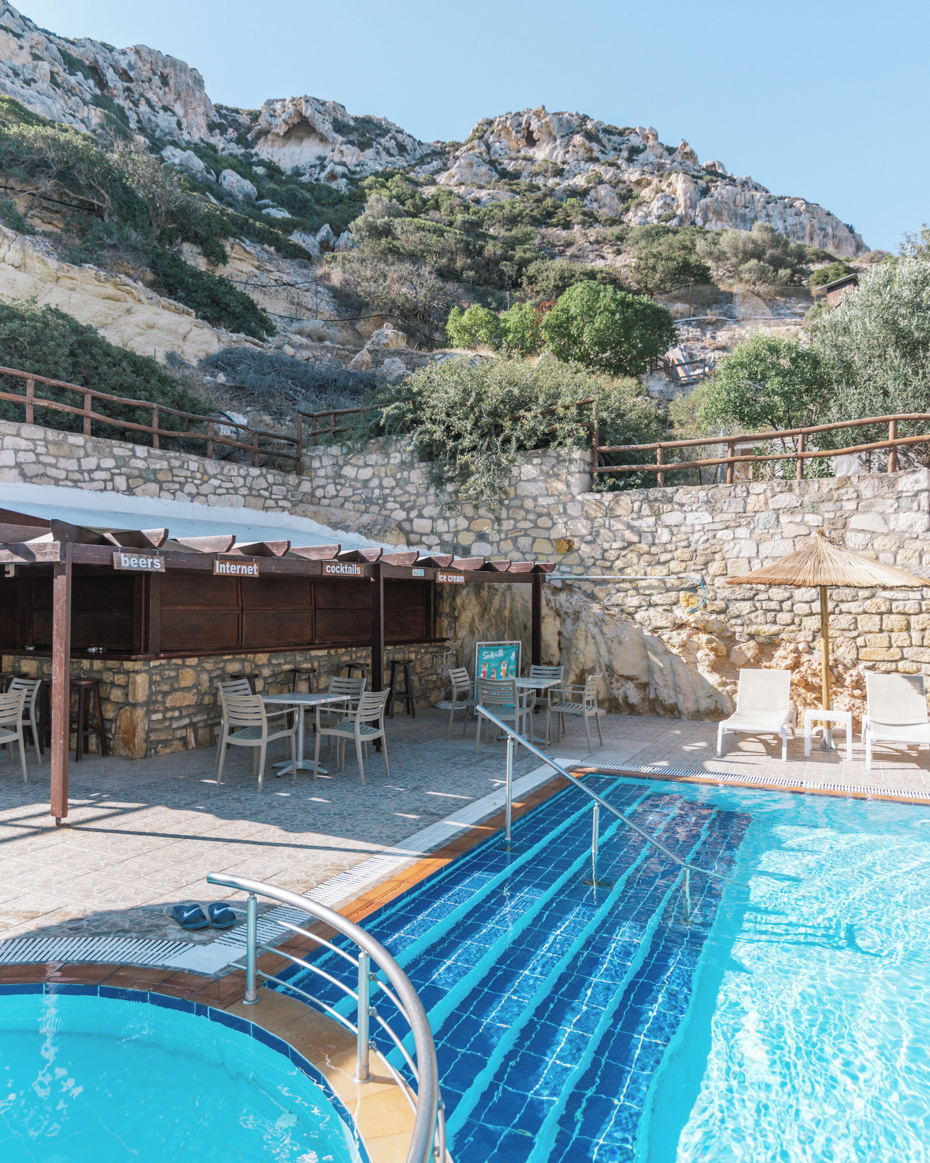 Ein Poolbereich mit einer Steinmauer, Liegestühlen und einer Bar, auf der es Schilder für Biere, Cocktails und Eis gibt.