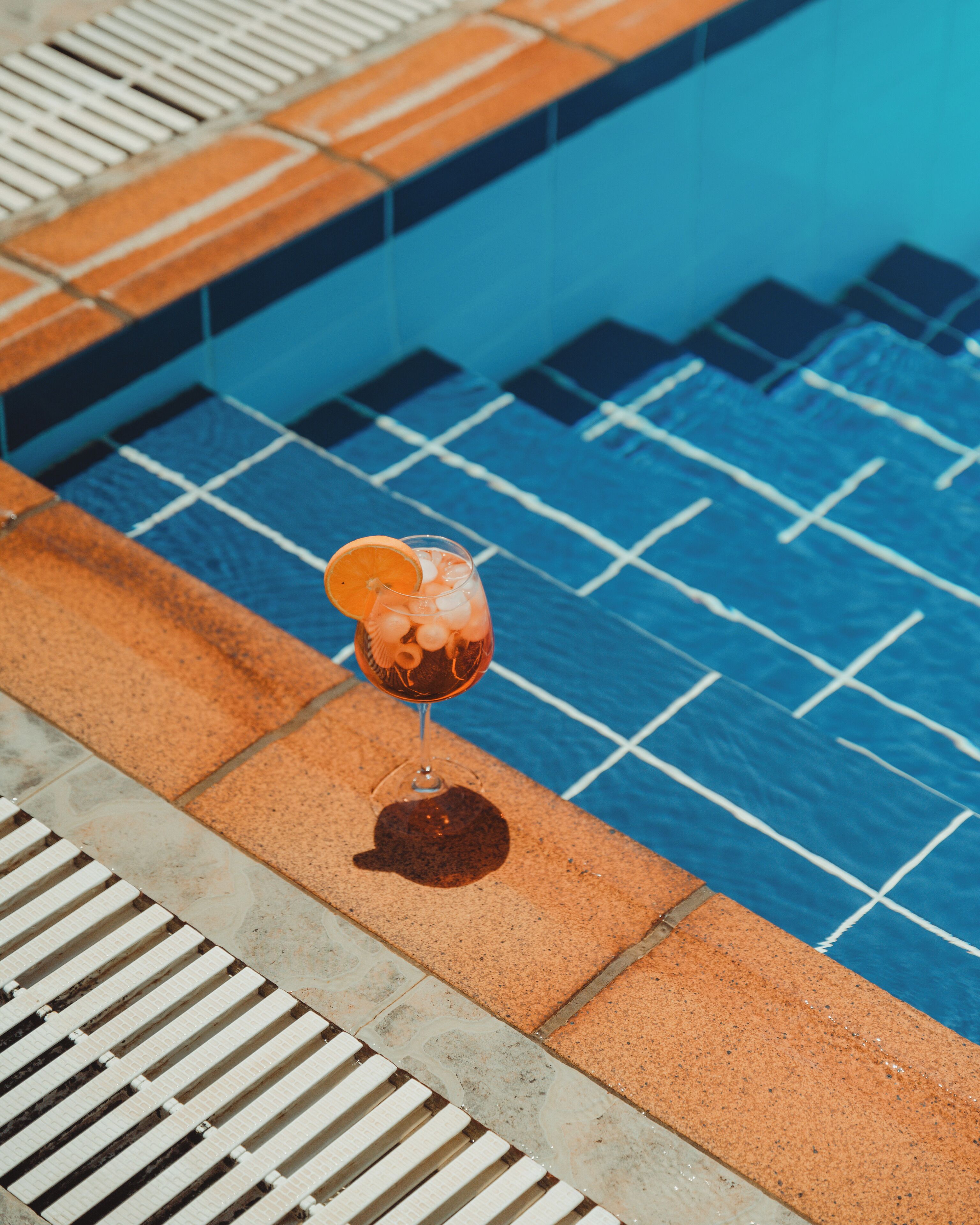 Un verre contenant une boisson, garni d’une tranche d’orange, posé au bord d’une piscine dont le fond est recouvert de carreaux bleus et qui est équipée d’un drain.