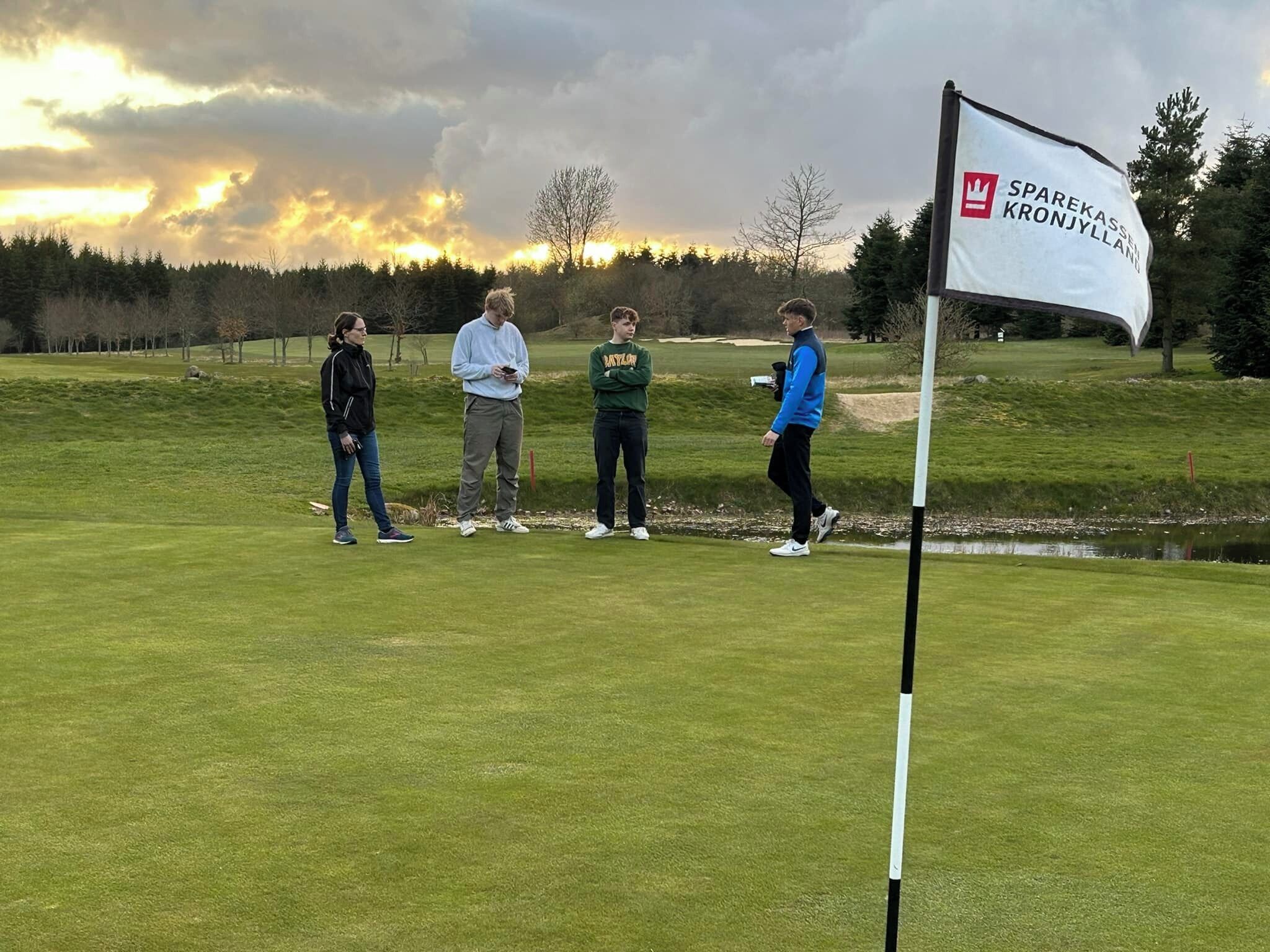 A golf course with a flag marked "SPAREKASSEN KRONJYLLAND" and four people standing on the green.