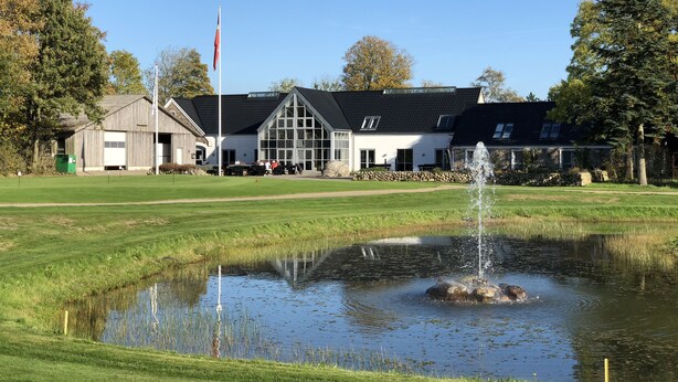A building with a large glass facade, a flagpole, and a pond with a fountain. - Tollundgaard Golf Park (Silkeborg)