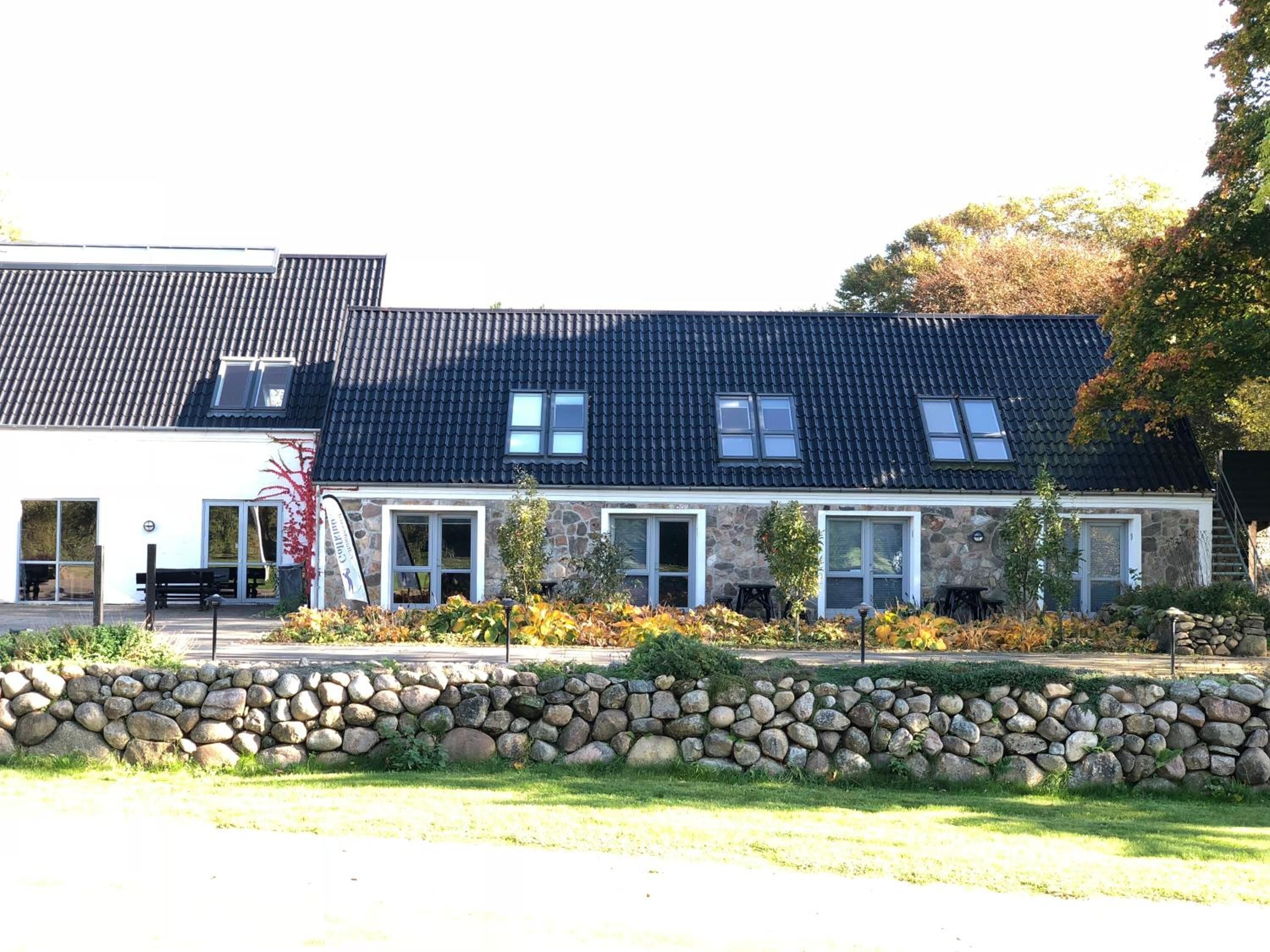 A two-story stone building with a dark tiled roof, stone walls, and a stone wall in the foreground.