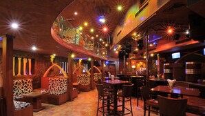 A restaurant with booths and bar seating, a high ceiling with hanging lights, and a bar area with a television. - Ramee California Juffair (Manama)