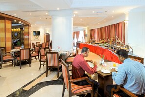 A hotel dining area with multiple tables and chairs, a buffet setup, and a large screen on the wall.