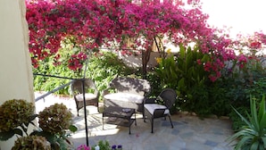 An outdoor patio with wicker furniture and a stone floor, surrounded by lush greenery and vibrant pink bougainvillea flowers. - Anniko (Chania)