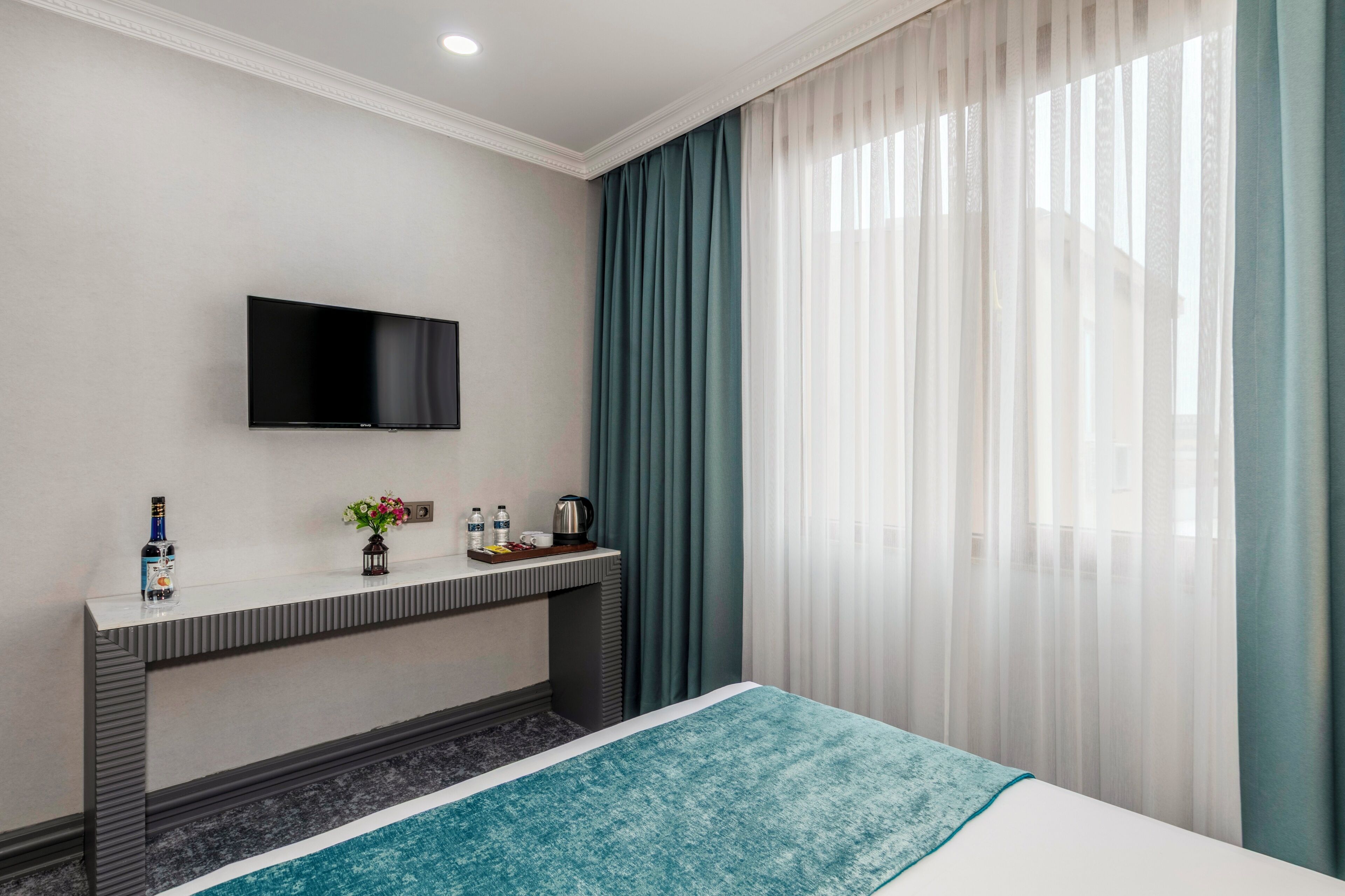 A hotel room with a bed, a TV mounted on the wall, a small table with a bottle, a vase with flowers, and a coffee maker.
