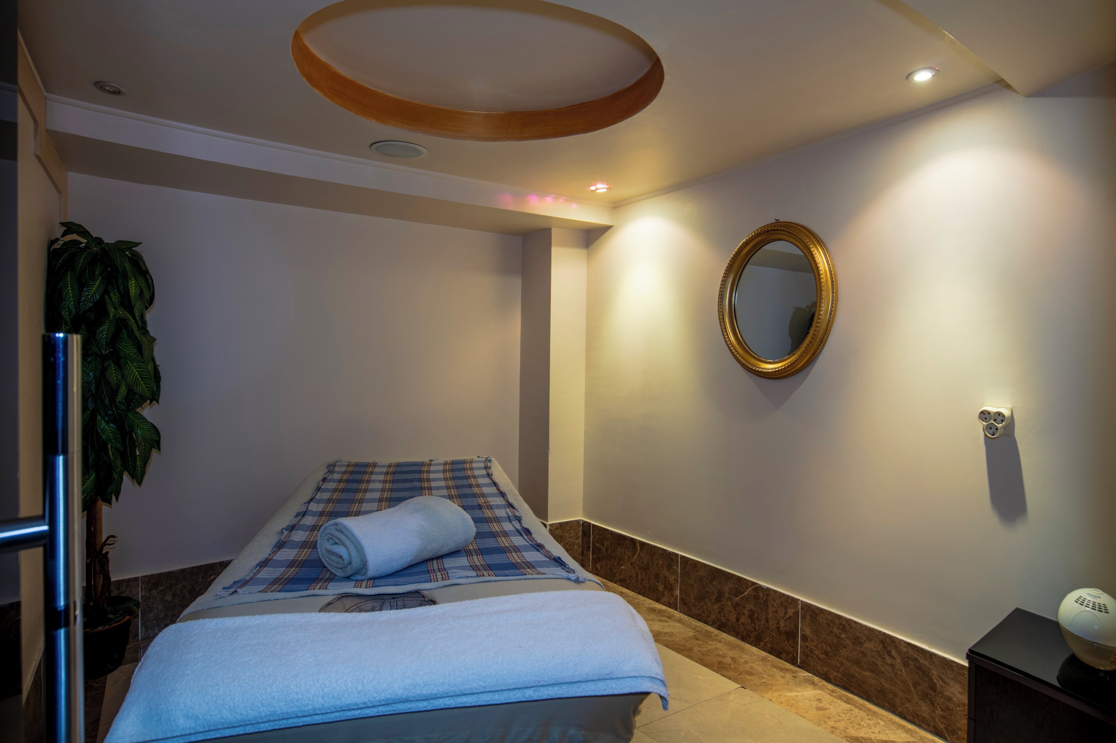 Habitación de hotel con cama, espejo en la pared, planta en maceta y una mesita de noche con lámpara.