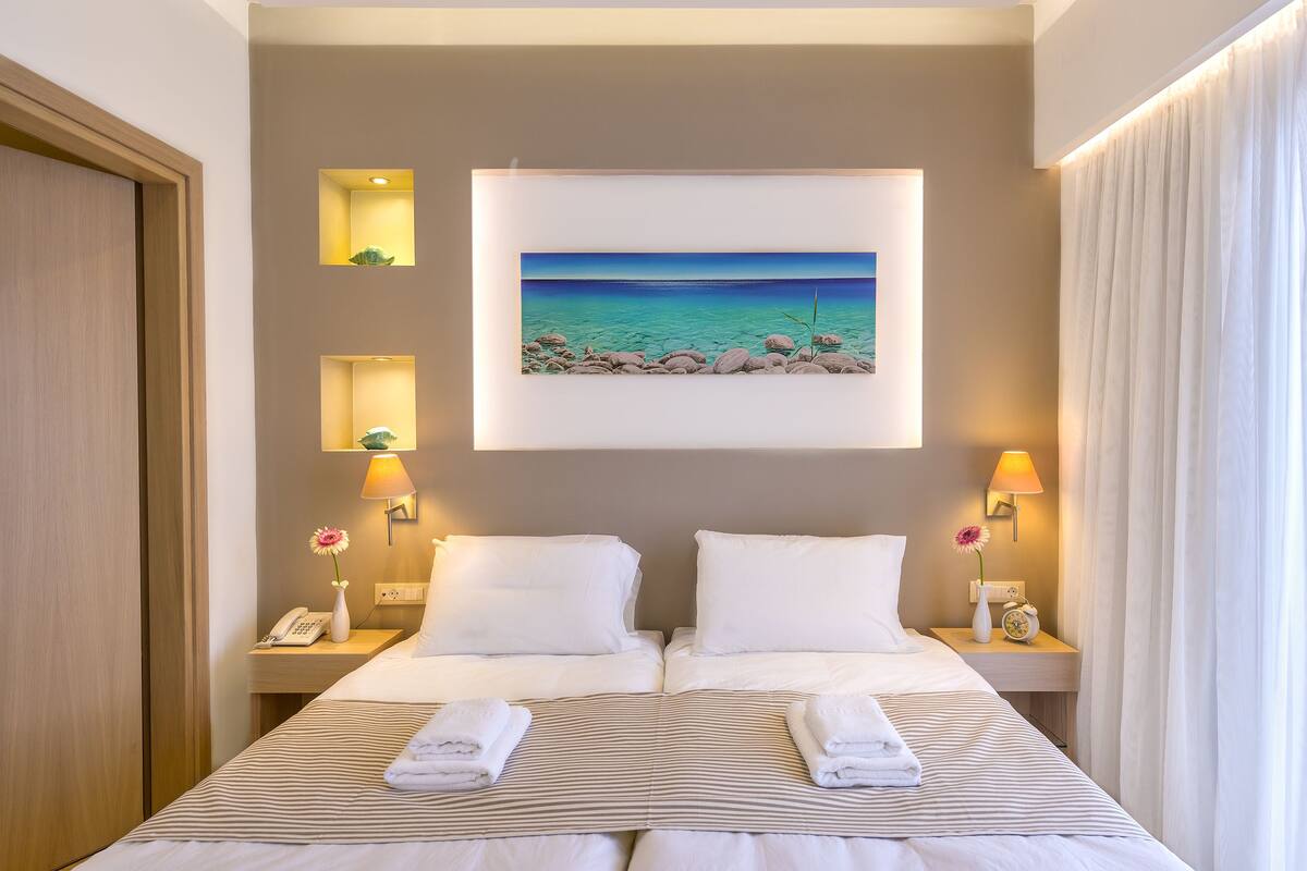 A hotel room with a bed, two bedside tables, a nightstand, a painting of a beach, and a window with curtains.