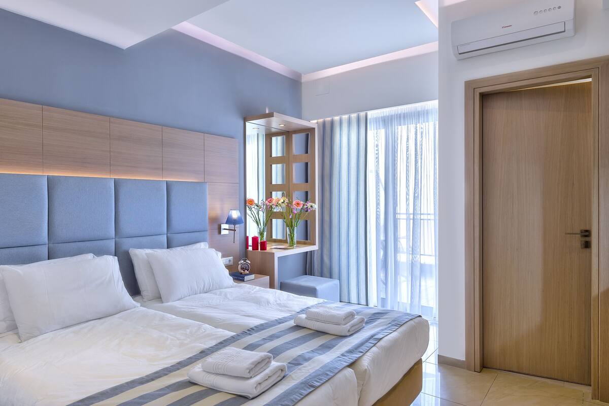 A modern hotel room with a large bed, a bedside table with a lamp, and a view of the outdoors through a window with curtains.