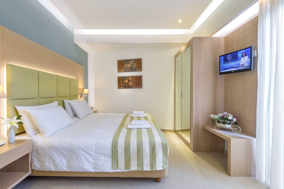 A modern hotel room with a large bed, bedside lamps, a flat-screen TV, and a small bench with flowers.