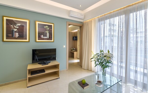 A modern living room with a flat-screen TV on a wooden stand, a glass coffee table with a book and an apple, and two framed floral paintings on the wall. - Leonidas Hotel & Apartments (Rethymno)