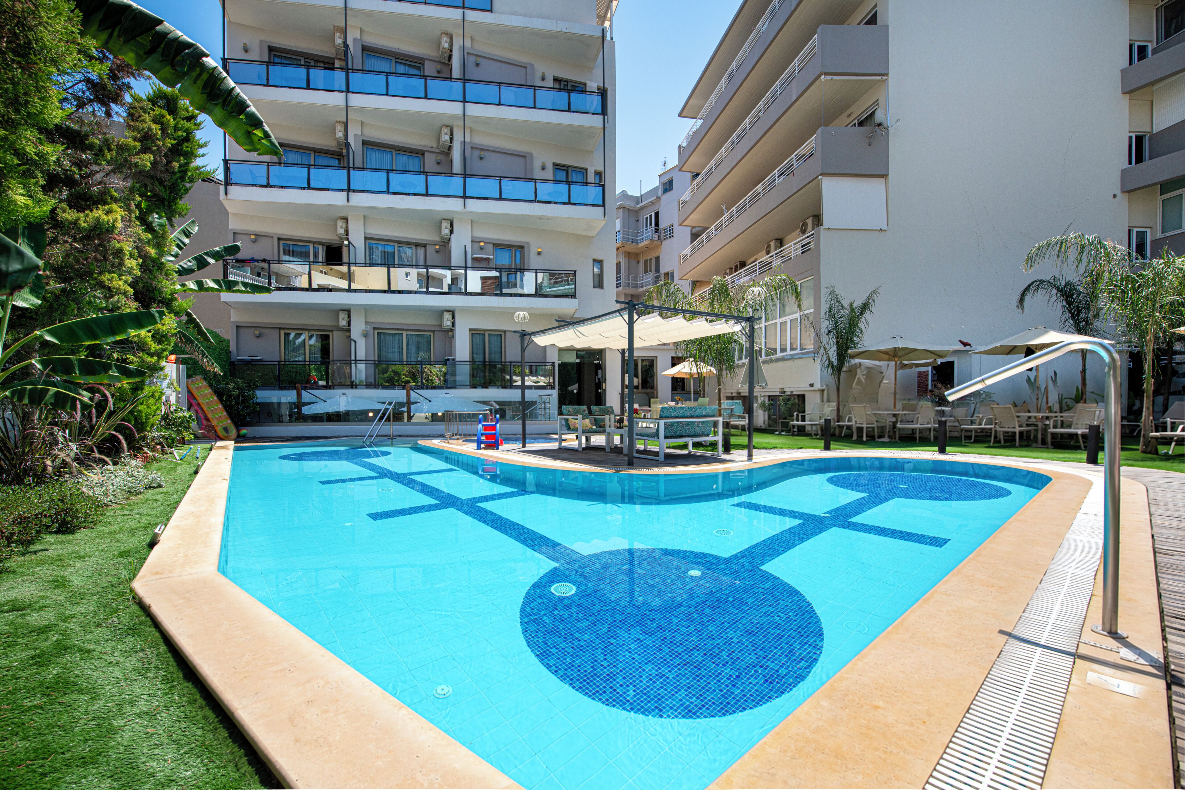 Una piscina con un diseño azul de mosaico, rodeada por un área con césped y un edificio de varios pisos con balcones.