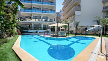 Una piscina con un diseño azul de mosaico, rodeada por un área con césped y un edificio de varios pisos con balcones.