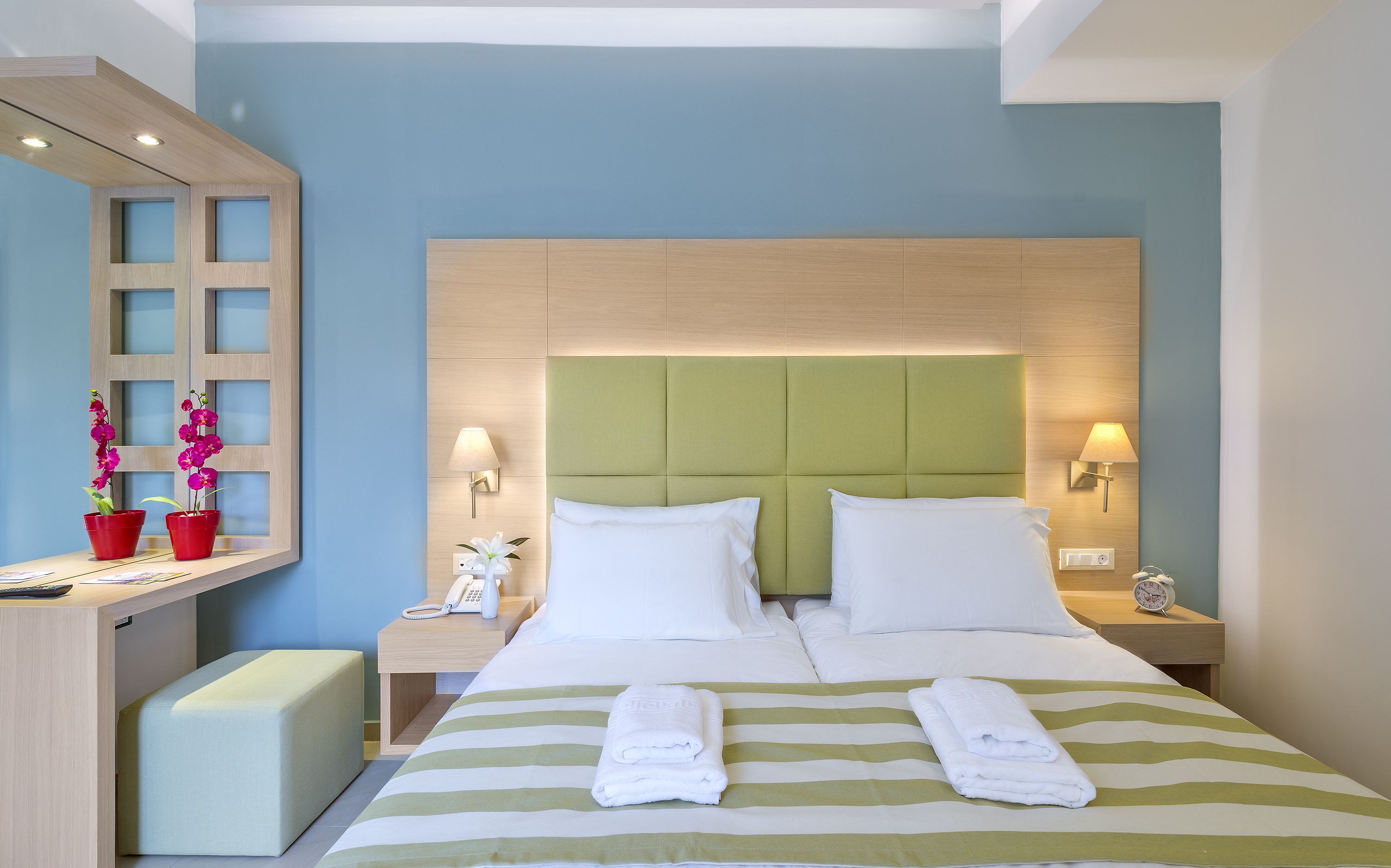 A hotel room with a bed, two bedside tables, a nightstand, a desk, and two potted orchids.