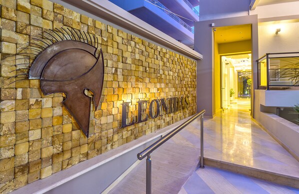A modern hotel entrance with a stone wall and a metal shield logo. - Leonidas Hotel & Apartments (Rethymno)