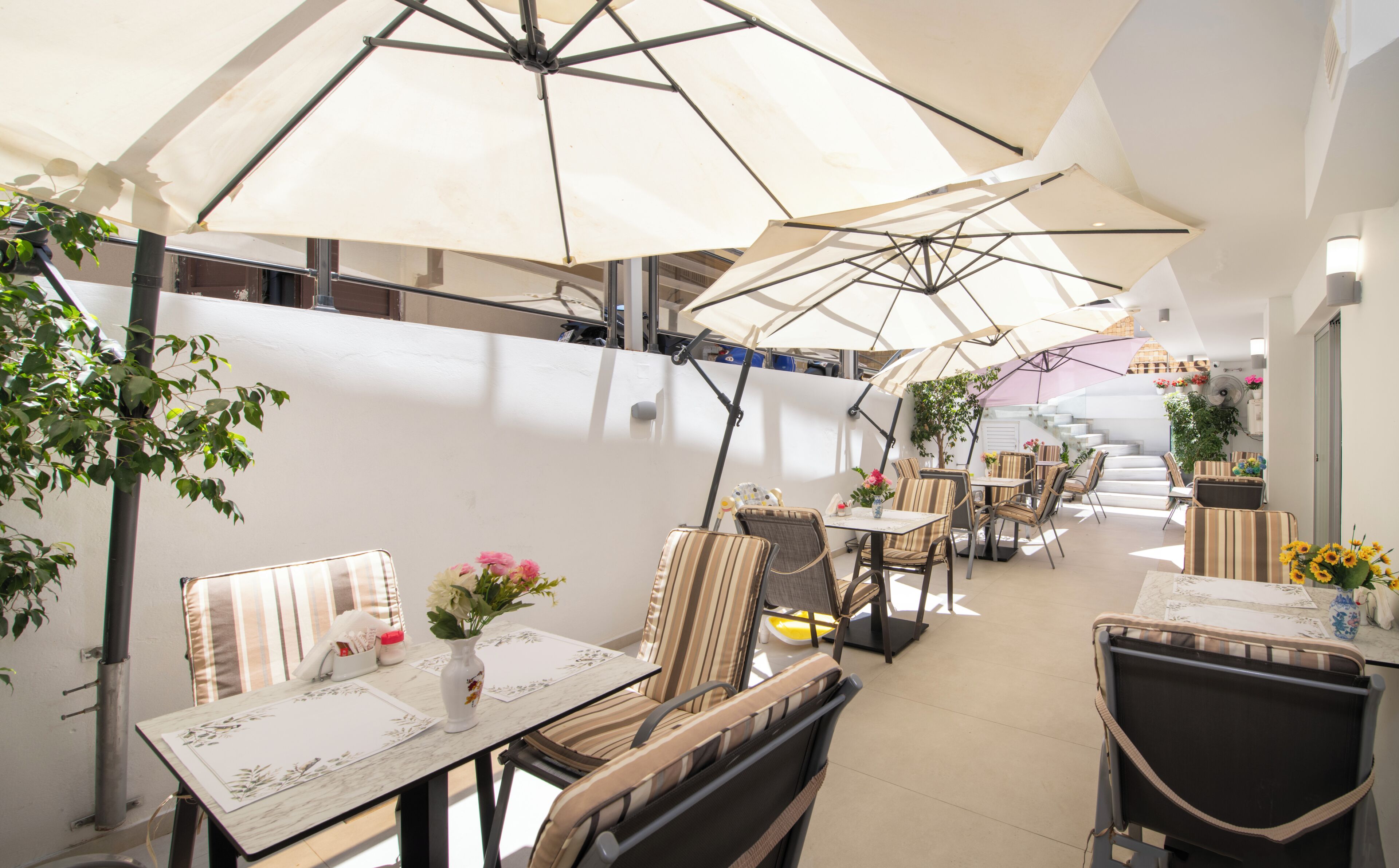 Un área de comedor al aire libre con varias mesas y sillas, protegida por grandes sombrillas y rodeada de plantas y una escalera.