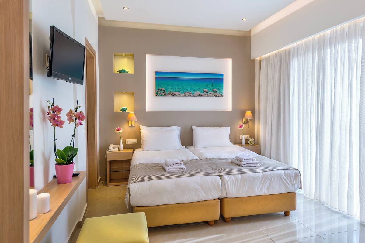 A hotel room with a large bed, a flat-screen TV, a painting of a beach, and a window with sheer curtains.
