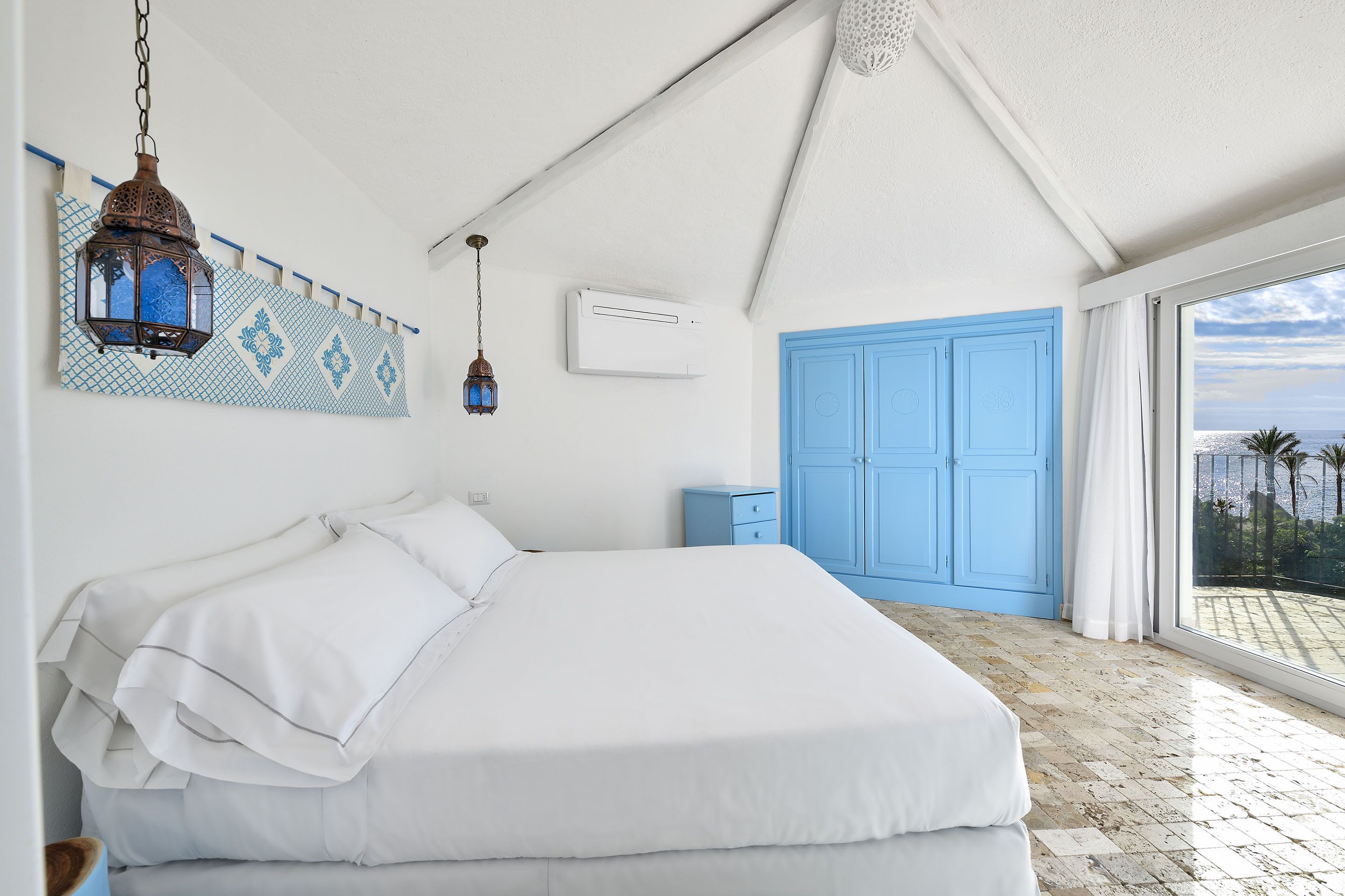 Une chambre avec un grand lit, une lanterne suspendue, une armoire bleue et une vue sur la mer par la fenêtre.