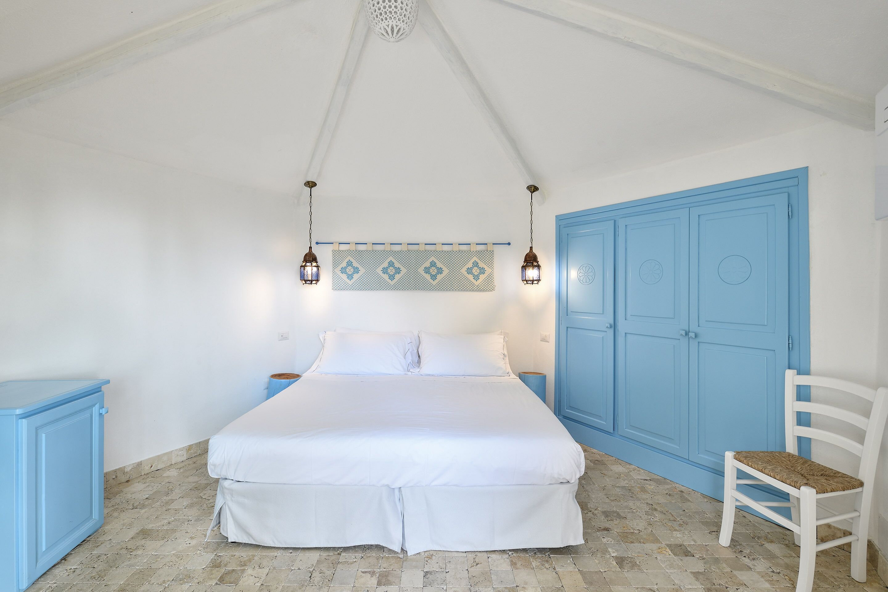 A bedroom with a bed, a blue wardrobe, a white chair, and a decorative wall hanging.