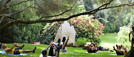 People lying on mats outdoors in a grassy area with trees and bushes in the background.