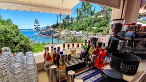 Zona de bar con vista a la costa rocosa y palmeras, con una variedad de botellas de licor y utensilios de bar.