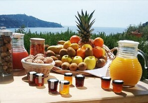 Una mesa con frutas variadas, una piña, frascos de conservas y una jarra de jugo de naranja, frente a un paisaje pintoresco.