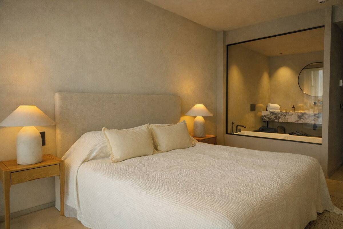 A hotel room with a large bed, two bedside tables with lamps, a large mirror, and a bathroom visible in the reflection.