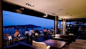 A modern lounge area with leather seating and a large window offering a view of a body of water and distant land. - Le Versailles (Villefranche-sur-Mer)