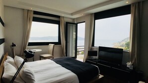 A hotel room with a bed, a TV, a desk, and a chair. There are large windows with curtains, and a view of the sea and mountains. - Le Versailles (Villefranche-sur-Mer)