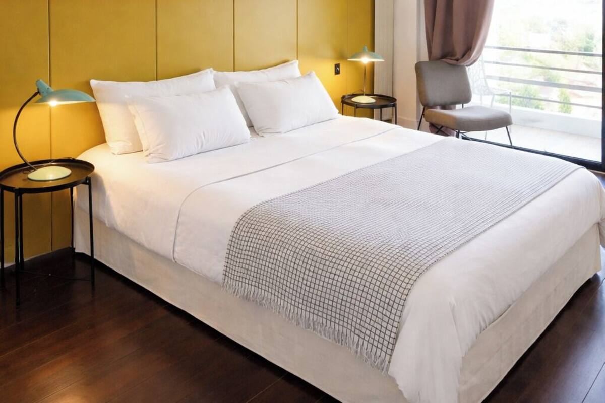 A hotel room with a large bed, two bedside tables with lamps, a chair, and a view of the outdoors through a window with curtains.