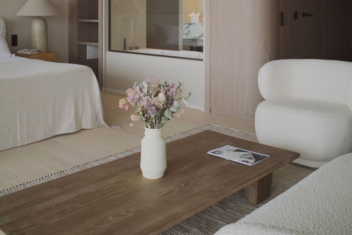 A modern hotel room with a bed, a white sofa, a wooden coffee table with a vase of flowers, a nightstand with a lamp, and a large sliding door closet.