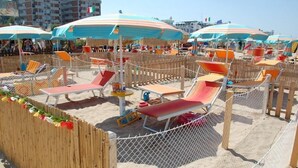 Zona de playa con tumbonas y sombrillas, rodeada por una valla de madera y una barrera de malla metálica.