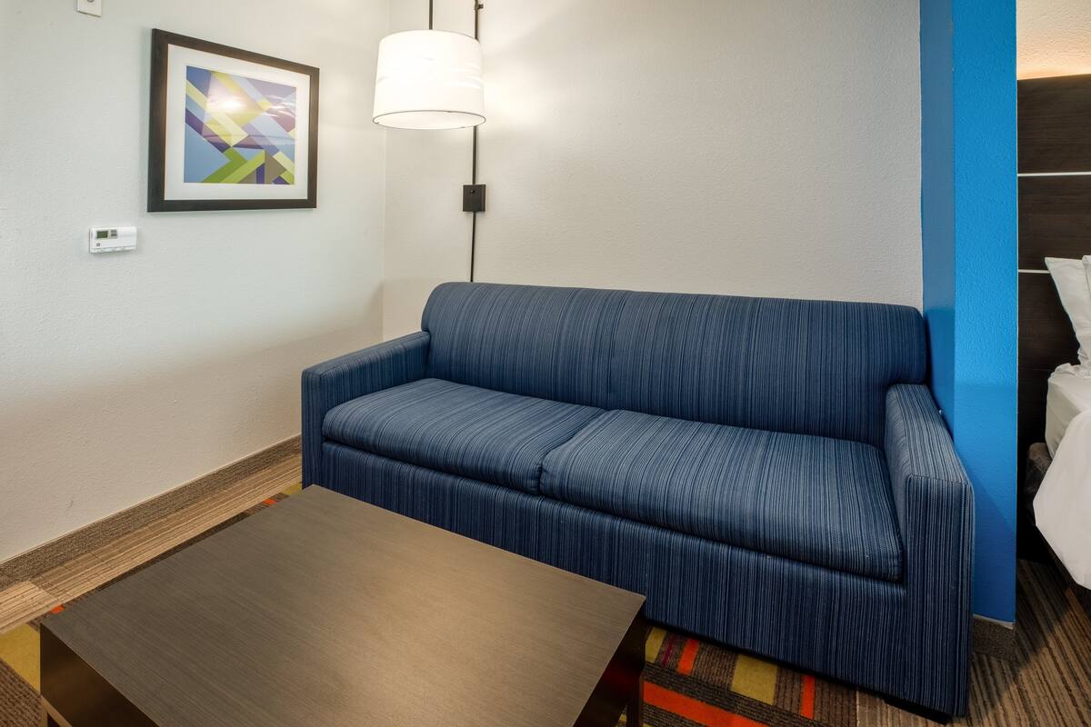 Habitación de hotel con sofá azul, una mesa de centro de madera, un cuadro abstracto enmarcado y una lámpara de pared.